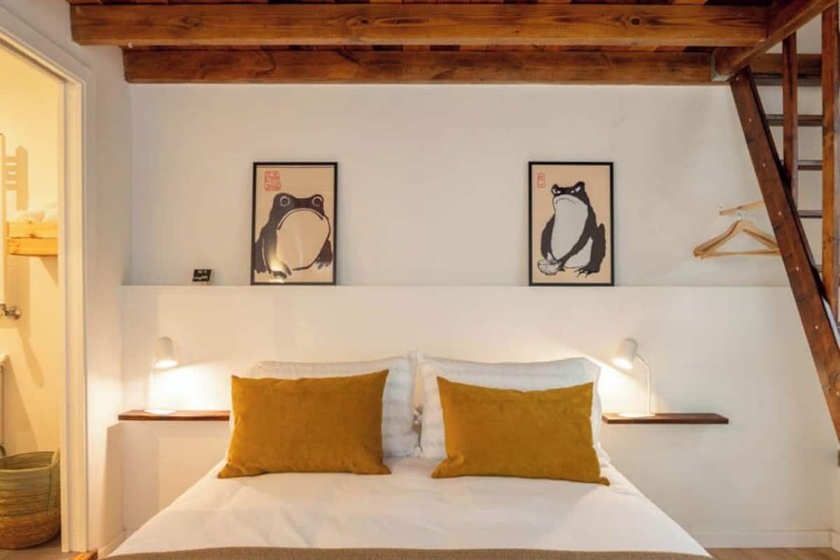 Un dormitorio con techo de madera, una cama con almohadas blancas y mostaza, dos cuadros enmarcados y lámparas de noche.