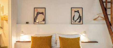 A bedroom with a wooden ceiling, a bed with white and mustard pillows, two framed wall art pieces, and bedside lamps.