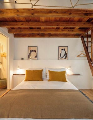 A bedroom with a wooden ceiling, a bed with white and mustard pillows, two framed wall art pieces, and bedside lamps.