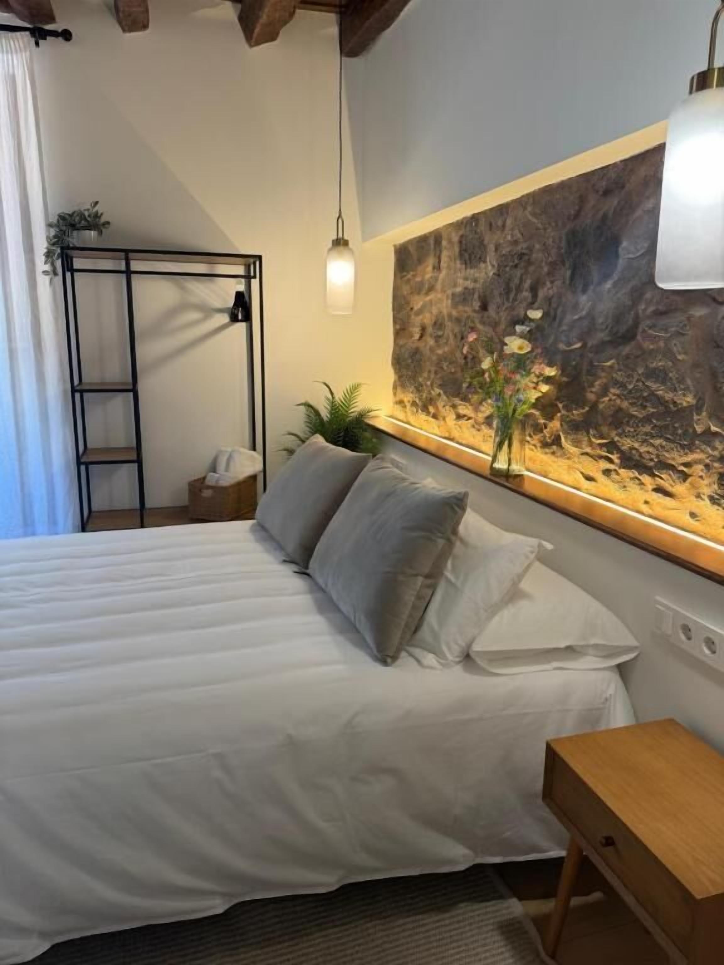 Een bed met witte beddengoed, een houten nachtkastje, een wandplank en een groot abstract schilderij.