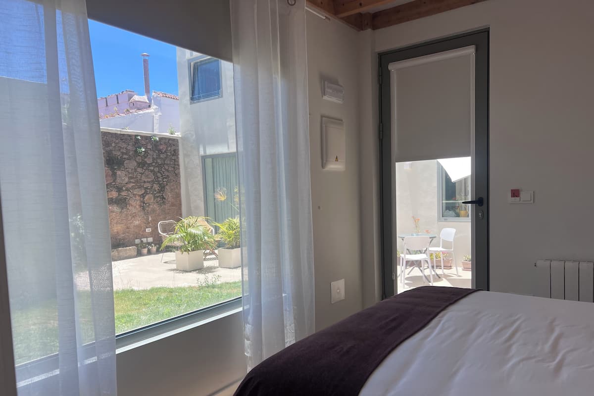 Un dormitorio con una cama, una ventana con cortinas y una puerta que da a un espacio exterior con sillas y plantas.