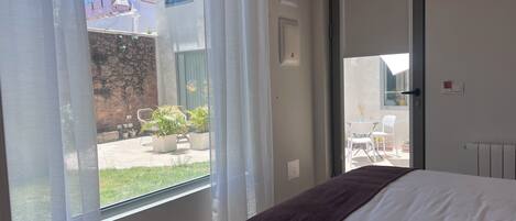 A bedroom with a bed, a window with curtains, a door leading to an outdoor area with chairs and plants.