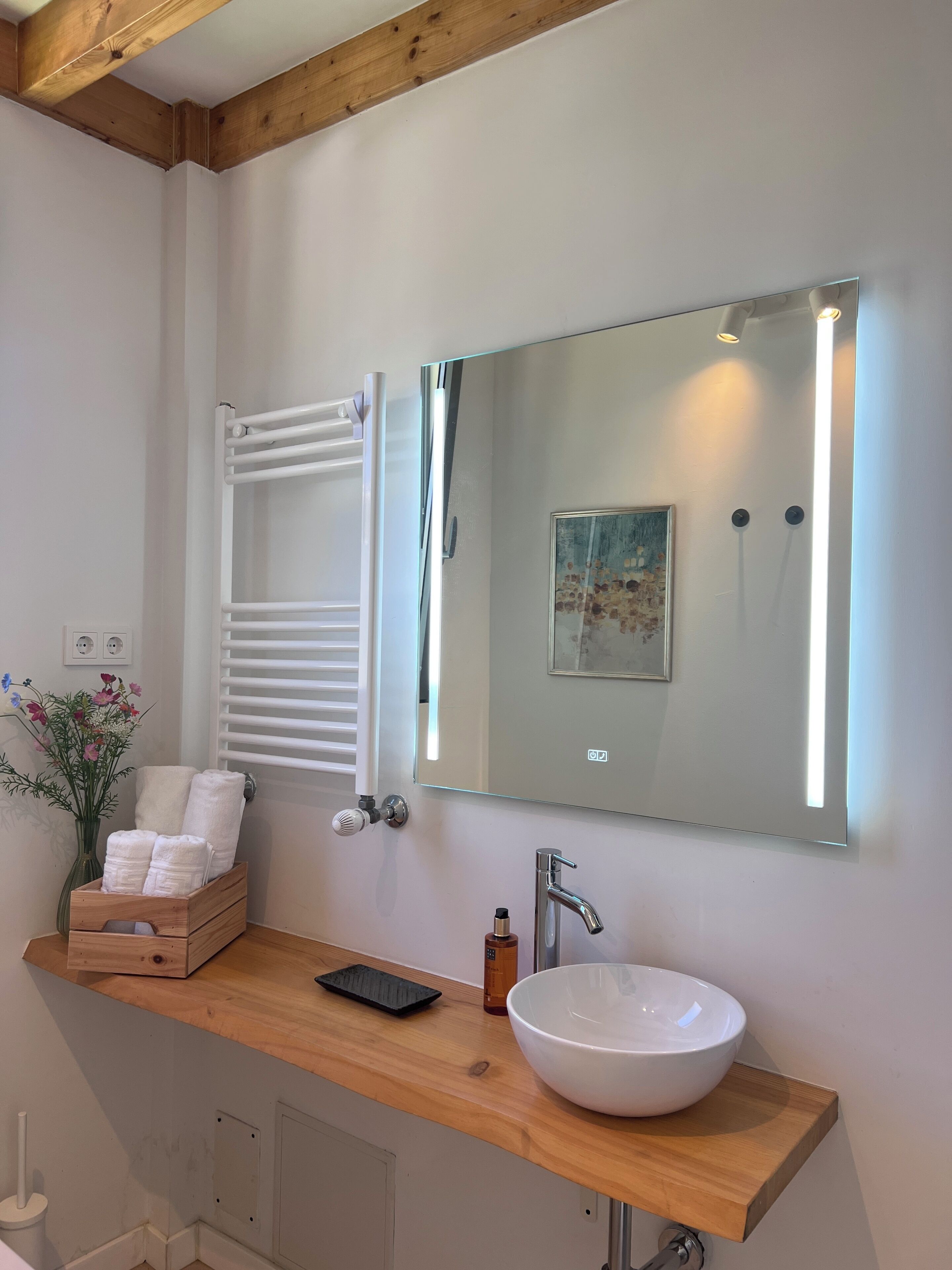 Een badkamer met een houten wastafelmeubel, een witte wastafel, een grote spiegel met verlichting, een handdoekhouder en een kleine vaas met bloemen.
