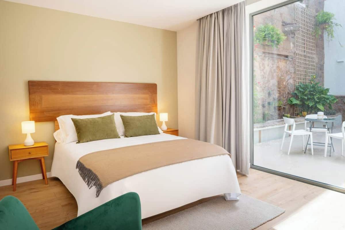Habitación de hotel con una cama grande, dos mesitas de noche con lámparas, vista a un patio exterior con sillas y una mesa, y cabecero de madera.