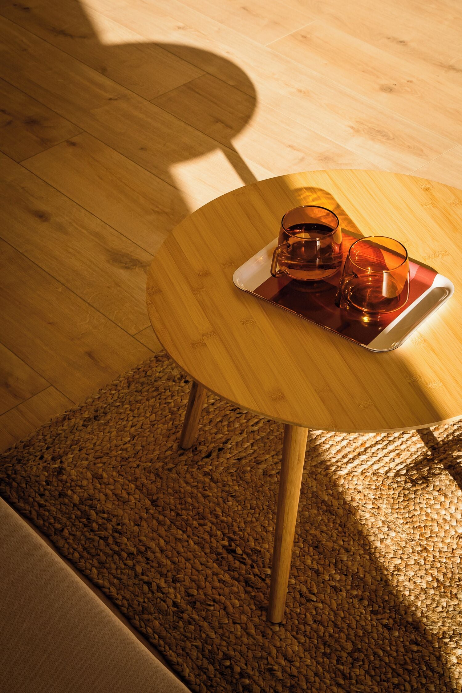 Een ronde houten tafel met een dienblad waarop twee glazen staan, een geweven vloerkleed en een houten vloer.