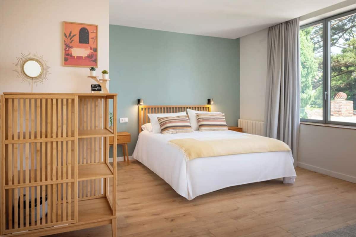 Un dormitorio con una cama, una estantería de madera, un espejo montado en la pared y una ventana con cortinas.