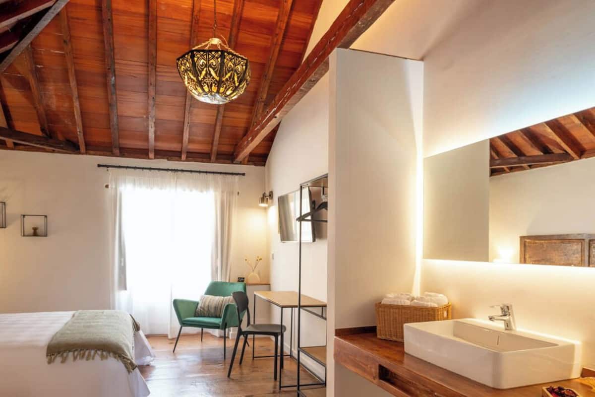Un dormitorio con cama, una silla, un escritorio y un baño con lavamanos y espejo.