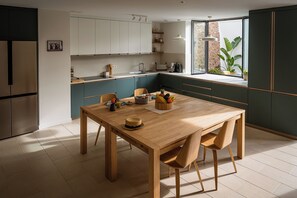 A modern kitchen with a wooden dining table, teal cabinetry, and a view of greenery outside.