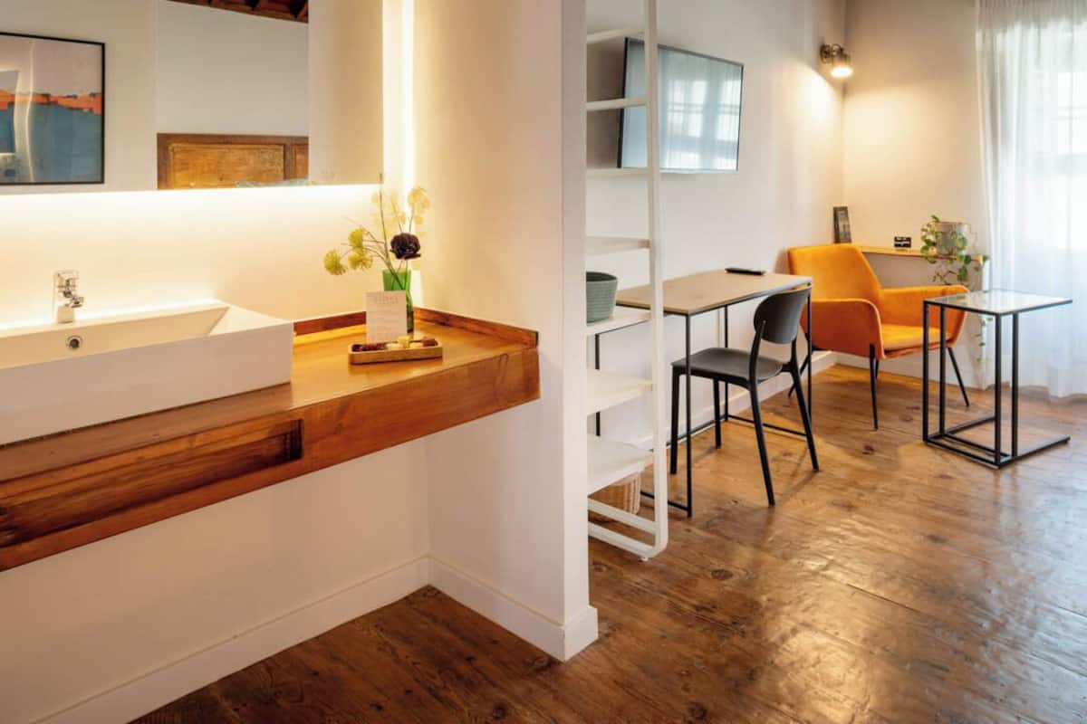 Un baño moderno con tocador de madera, un lavamanos blanco y un espejo. Hay una zona de comedor con mesa, sillas y vistas a la ciudad.