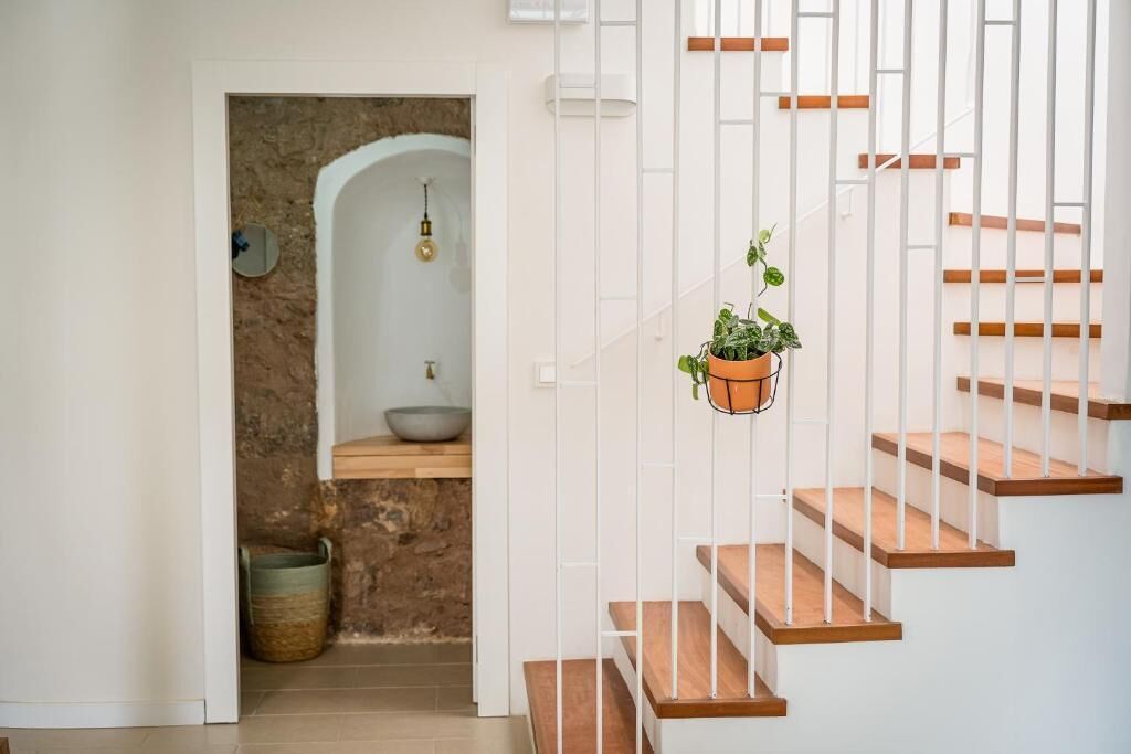 Un escalier avec des marches en bois et des rampes blanches, une plante suspendue et un mur de pierre orné d’un récipient et d’un luminaire.
