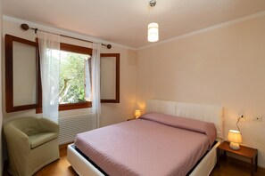Ein Schlafzimmer mit einem Bett, einem Sessel, einem Nachttisch mit Lampe, einem Fenster mit Vorhängen und einer Heizung.