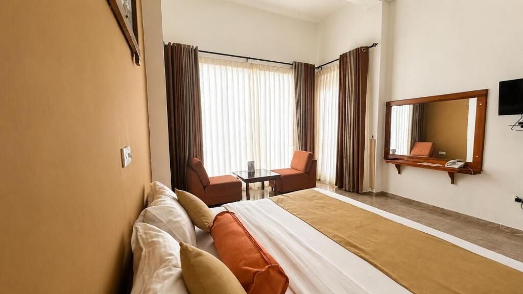Quarto de hotel com uma cama, duas cadeiras, uma mesa, uma televisão e um espelho grande.