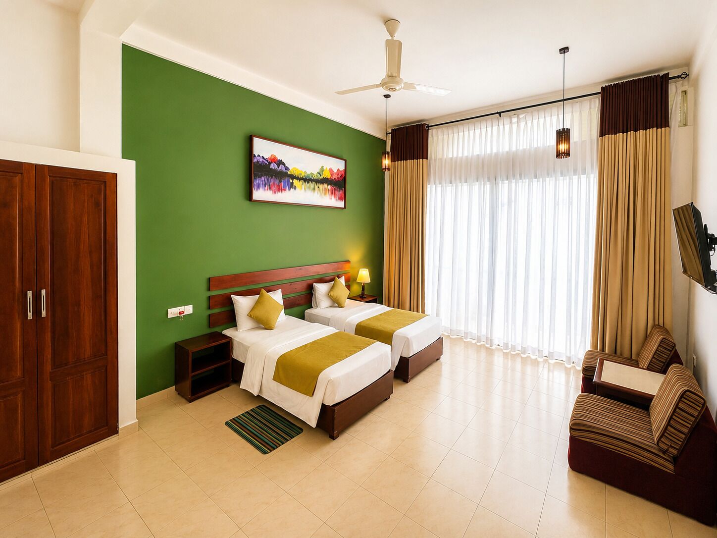 Une chambre d’hôtel avec deux lits, un mur d’accent vert, un ventilateur de plafond, une grande fenêtre avec des rideaux et un canapé.