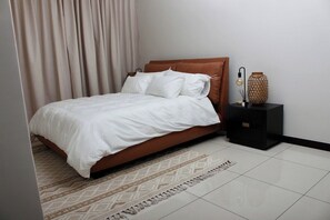 A neatly made bed with white bedding, a brown headboard, a patterned rug, a black nightstand with a vase and a lamp, and a curtain. - The Cozy Maison (Midrand)
