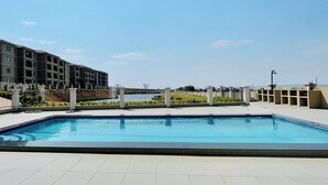 Una piscina rectangular con solárium alicatado, rodeada por una cerca baja, y contigua a un edificio de varios pisos con balcones.