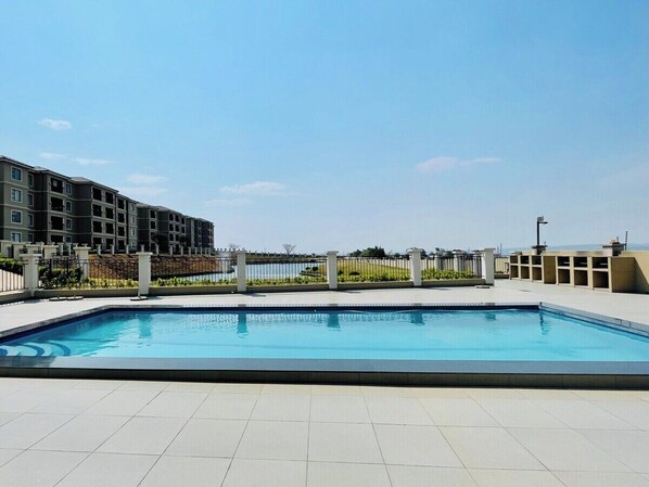 A rectangular swimming pool with a tiled deck, surrounded by a low fence, and adjacent to a multi-story building with balconies. - The Cozy Maison (Midrand)