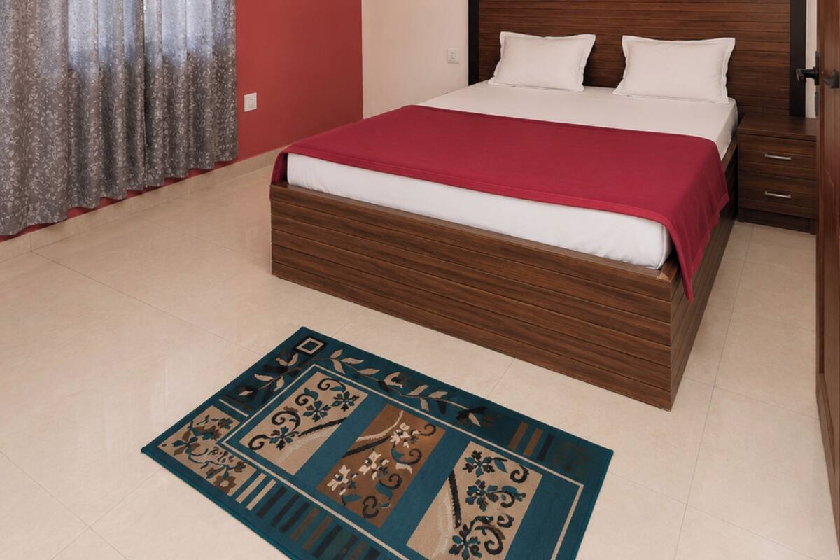 A bedroom with a bed, bedside tables, an air conditioning unit, a ceiling fan, a decorative rug, and a painting on the wall.