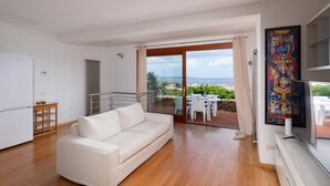 A modern living room with a white sofa, a dining area with a table and chairs, a TV, and a view of the sea. - Casa Ald a Manna by Wonderful Italy (Olbia)