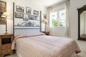 Ein Schlafzimmer mit einem Bett, zwei Nachttischen, einem Spiegel, einem Fenster mit Vorhängen und einer Wand mit gerahmten Bildern.