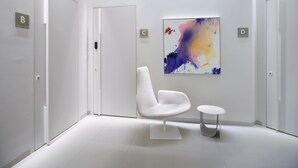 Una habitación moderna y minimalista con paredes blancas, un cuadro abstracto colorido, una silla blanca y una pequeña mesa redonda.