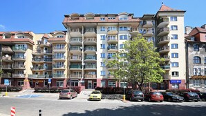 Un edificio residencial de varios pisos con balcones, un estacionamiento y un árbol en la fachada.