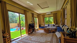 A spacious living room with a large window, a sofa, a television, and a small table. - Perfect 2-bedroom Villa with Pool in delightful Ourika (Ourika)