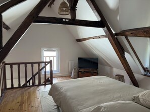Ein Schlafzimmer mit geneigter Decke, Holzbalken, einem Bett, einem Fernseher auf einer Holzkommode und einem Fenster.