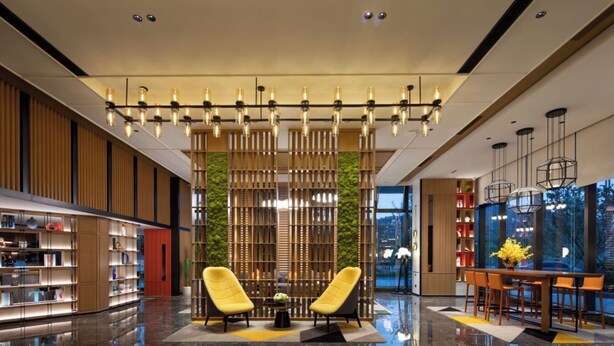 A modern lobby with a reflective marble floor, two yellow armchairs, a wooden partition with vertical slats, and a dining area with a table and chairs. - Hampton by Hilton Huzhou Deqing (Huzhou)