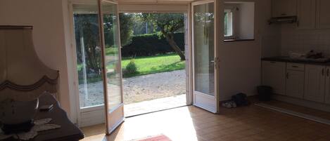 Une pièce dotée d’une porte-fenêtre coulissante donnant sur un jardin, d’un coin cuisine avec des placards et d’une table à manger avec une lampe.