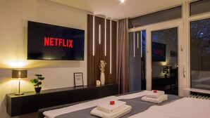 A modern hotel room with a large flat-screen TV displaying 'NETFLIX,' a bedside table with a lamp, a small potted plant, and neatly folded towels on the bed. - Design-FeWo at the Möhnesee with unique lake view, balcony, pool, sauna, elevator (Möhnesee)