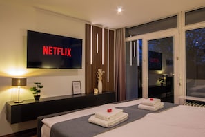 A modern hotel room with a large flat-screen TV displaying 'NETFLIX,' a bedside table with a lamp, a small potted plant, and neatly folded towels on the bed. - Design-FeWo at the Möhnesee with unique lake view, balcony, pool, sauna, elevator (Möhnesee)