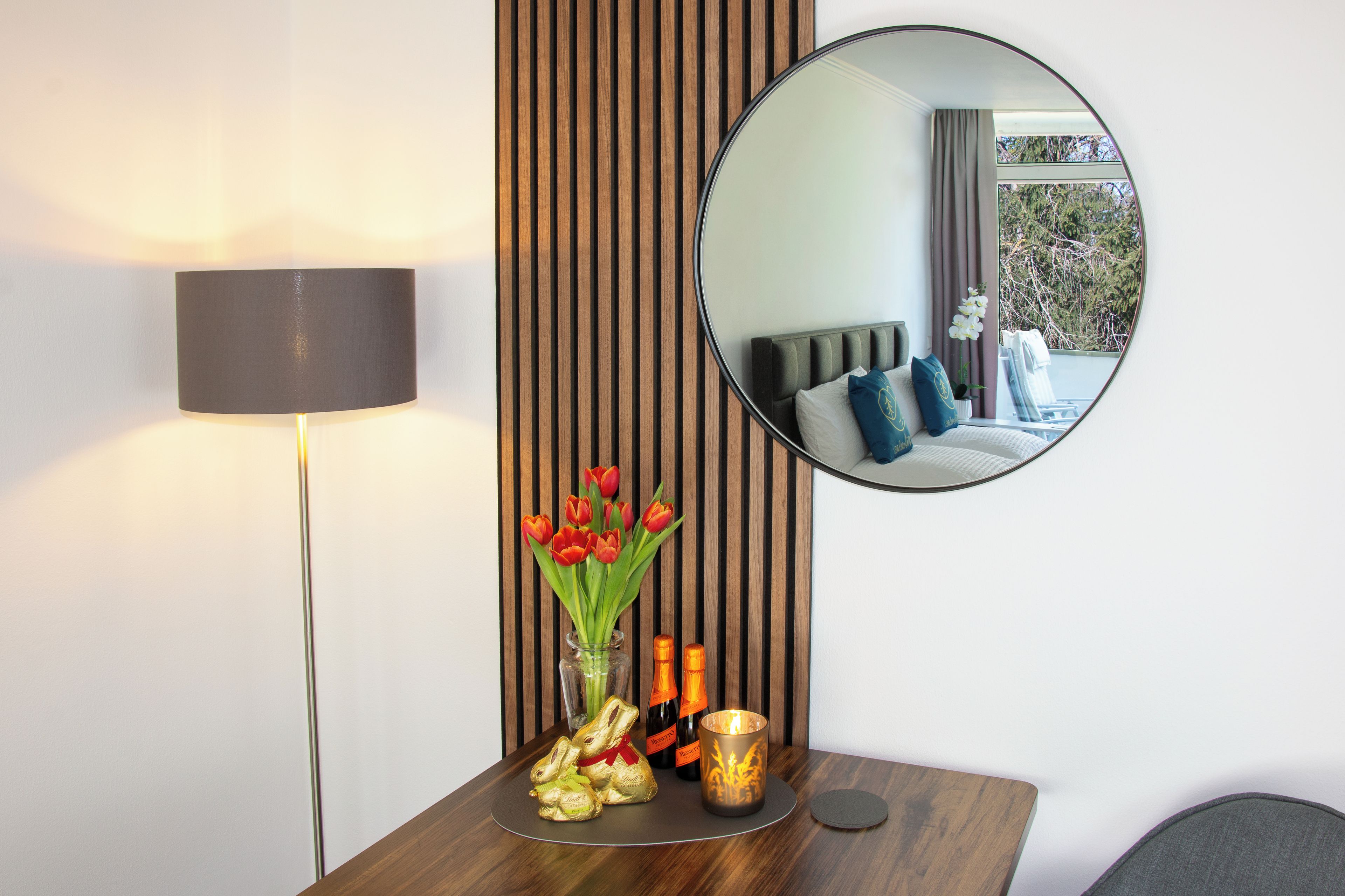 Une chambre d’hôtel avec un mur lambrissé en bois, un miroir rond, une table de chevet avec une lampe, un vase de tulipes et une bouteille de champagne.