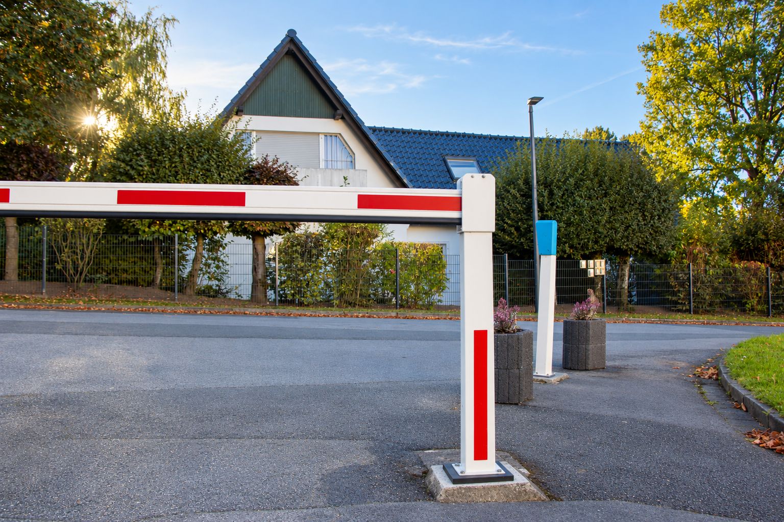 Een toegangspoort met een rode en witte barrière, een lantaarnpaal en een gebouw met een blauw dak op de achtergrond.