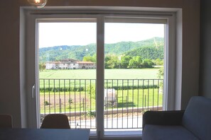 A room with a sliding glass door leading to a balcony with a view of a green field and a building in the distance. - Mamì cottage: accommodation Bisalta (Grassini)