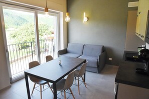 A modern kitchen with a dining area, a grey sofa, and a balcony with a view of greenery. - Mamì cottage: accommodation Bisalta (Grassini)