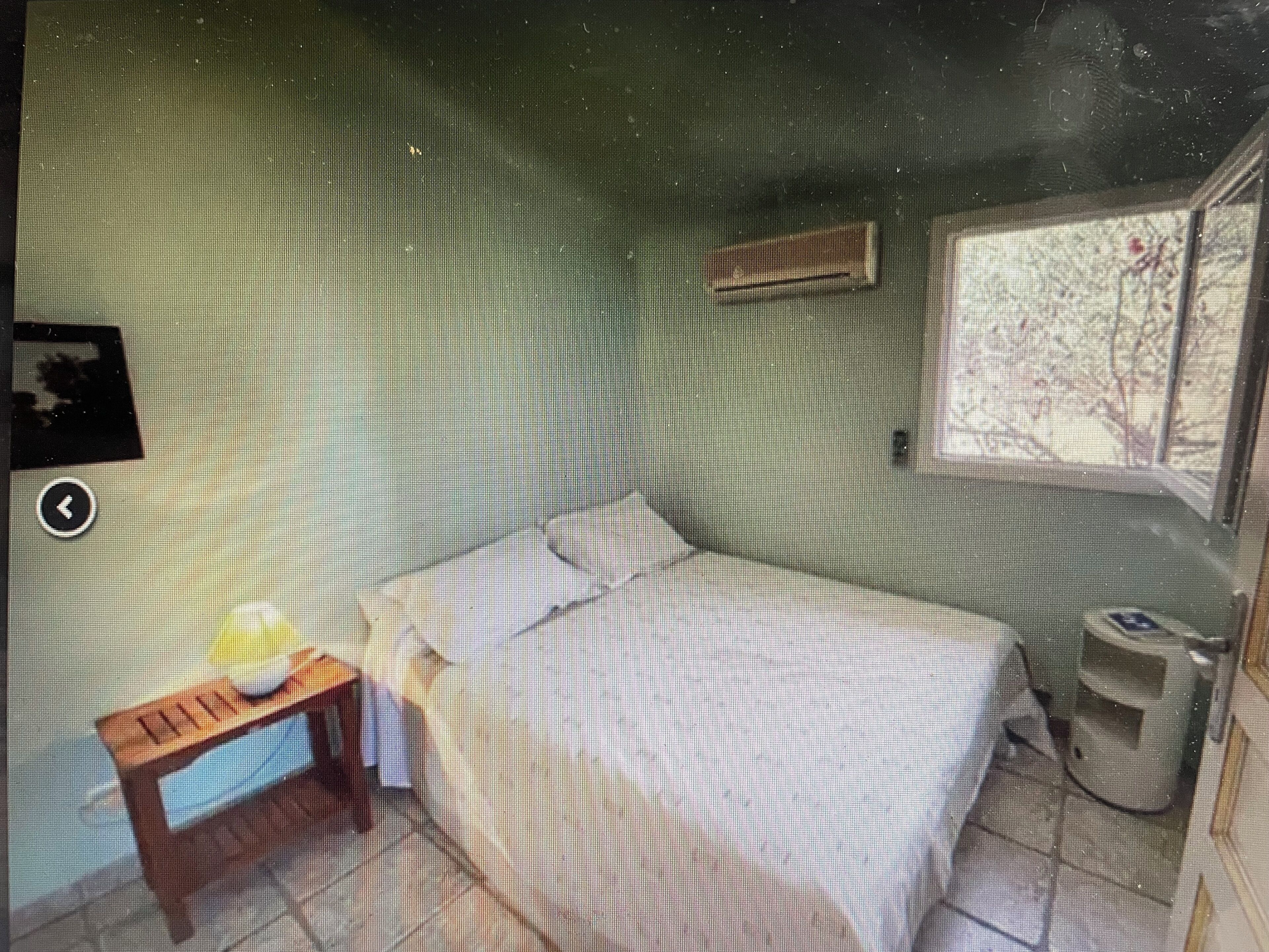 Une chambre à coucher avec un lit, une petite table avec une lampe, une fenêtre donnant sur des arbres et un climatiseur fixé au mur.