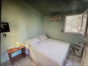 Une chambre à coucher avec un lit, une petite table avec une lampe, une fenêtre donnant sur des arbres et un climatiseur fixé au mur.