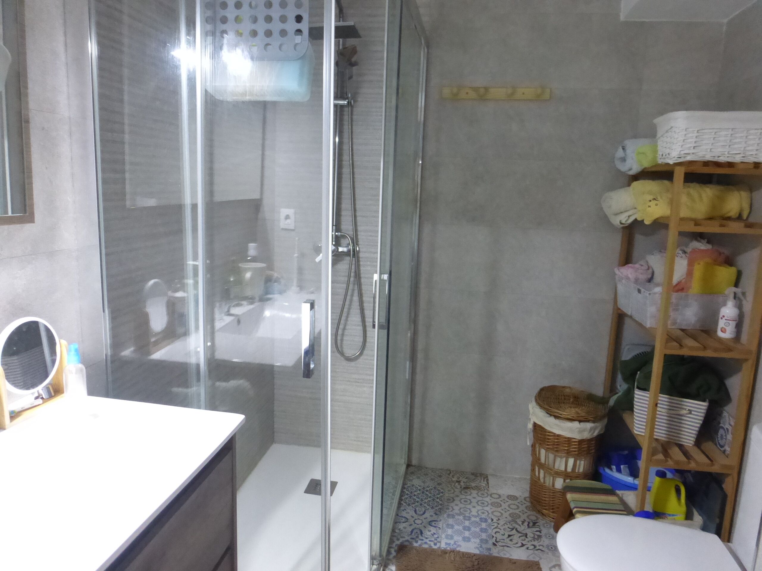 Un baño con cabina de ducha de vidrio, un lavabo blanco y un estante de madera con artículos de aseo.
