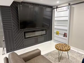 Ein modernes Wohnzimmer mit einem großen Flachbildfernseher an einer dunklen Wand, einem runden Holztisch, einem Ledersofa und einer weißen Bücherreol.