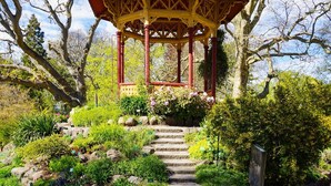 Ein Gartenpavillon mit roter und gelber Struktur, umgeben von einem Garten mit verschiedenen Pflanzen und einer Steintreppe, die zu ihm hinaufführt.