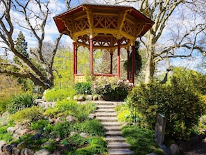 Ein Gartenpavillon mit roter und gelber Struktur, umgeben von einem Garten mit verschiedenen Pflanzen und einer Steintreppe, die zu ihm hinaufführt.