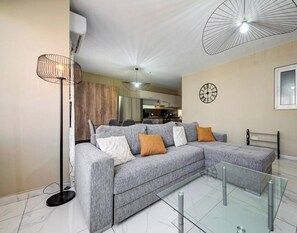 A modern living room with a grey sectional sofa, a floor lamp, a glass coffee table, and a kitchen area in the background. - Modern Central 3BR Apartment w/ WiFi, AC & Balcony (San Ġwann)