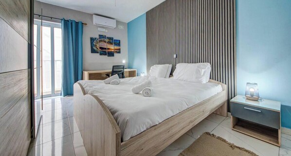 A modern bedroom with a wooden bed, a desk, a chair, and a wall-mounted air conditioner. - Modern Central 3BR Apartment w/ WiFi, AC & Balcony (San Ġwann)