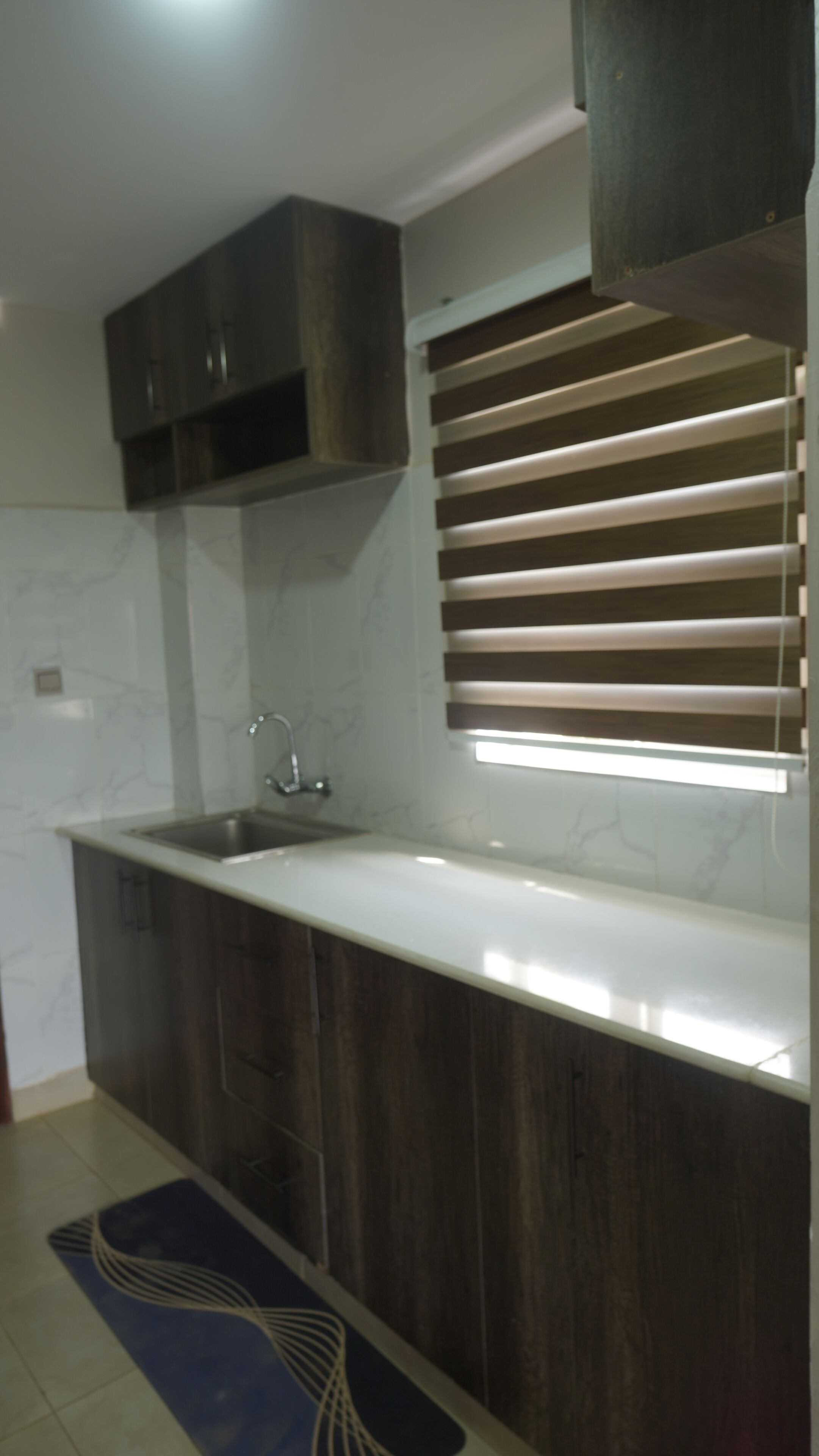 A kitchen with a white countertop, dark wooden cabinets, a stainless steel sink, and a window with blinds.