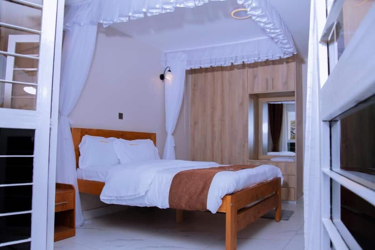 A bedroom with a wooden bed, white bedding, a nightstand, a wooden wardrobe, and a window with curtains.