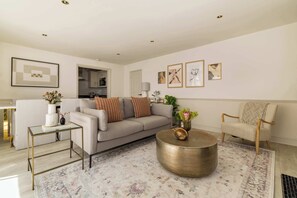 A living room with a sofa, coffee table, and armchair, a dining area with a table and chairs, and a kitchen visible through an open doorway. - Brass & Shaker Clapham (London)