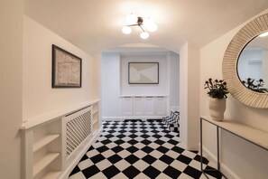 A hallway with a black and white checkered floor, white walls, a framed artwork, a ceiling light fixture, and a round mirror. - Brass & Shaker Clapham (London)