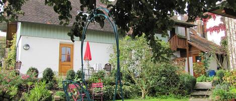 A garden with a swing chair, a white house, and a red umbrella.