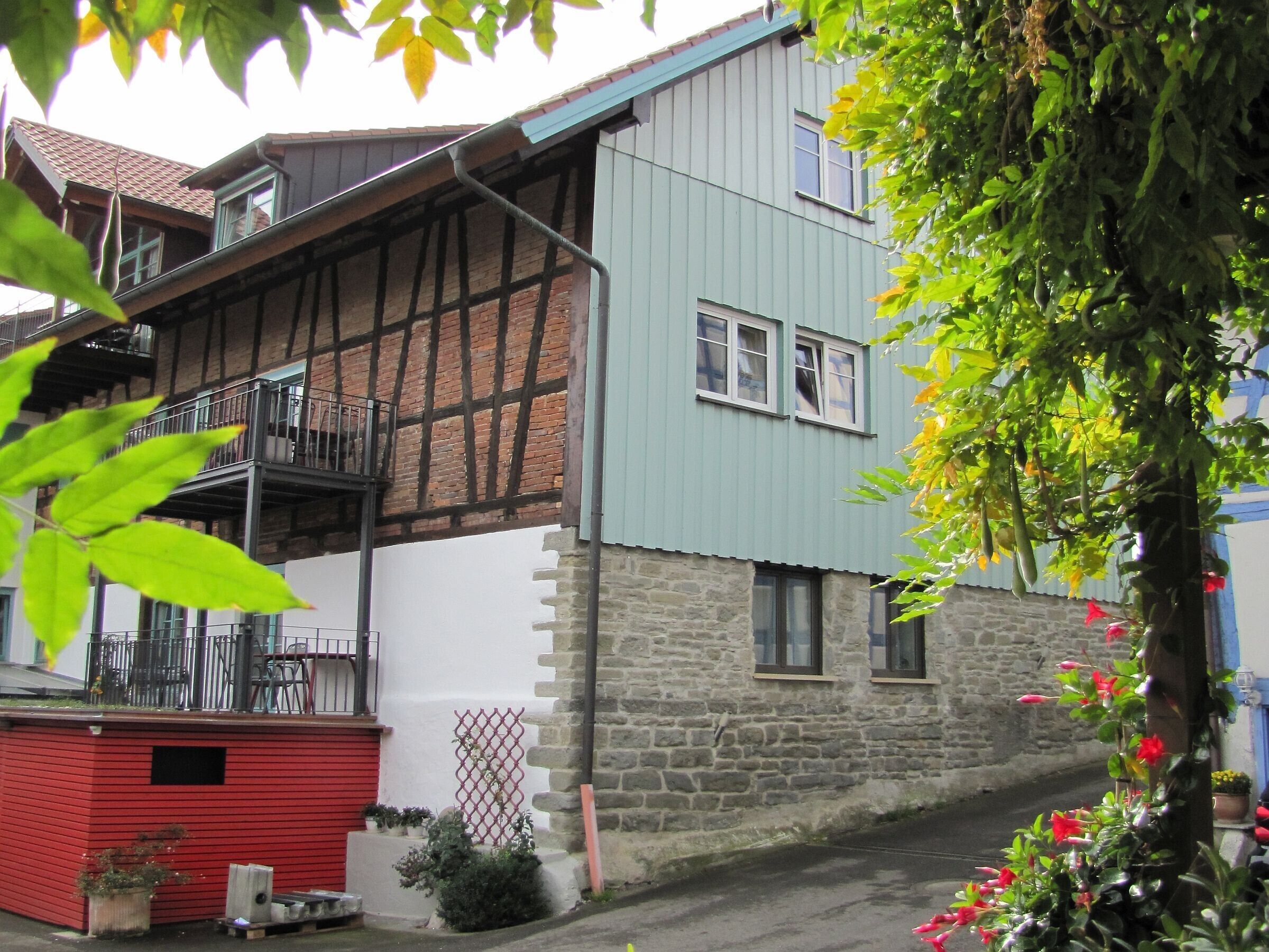 A two-story building with a mix of exposed timber framing and stone walls, a small red shed, and a balcony with metal railings.