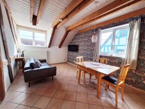 Ein gemütliches Dachzimmer mit Holzbalkendecke, einem schwarzen Ledersofa, einem Holzesstisch mit Stühlen und einem an der Wand befestigten Fernseher.